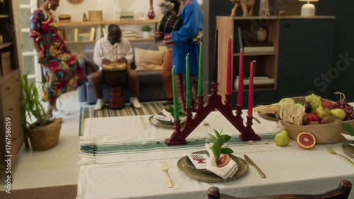Table set with white cloth, plates, cutlery, basket of fresh fruits and candleholder with red and green candles in foreground, Black friends playing goblet drums and dancing in background