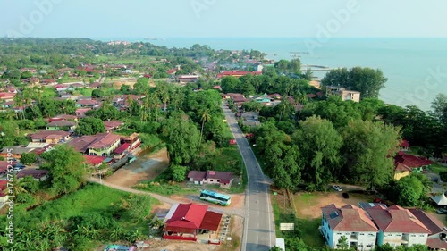 Aerial view of a coastal village in Malaysia with houses, greenery, and a road leading to the beach, showing peaceful rural lifestyle by the sea.