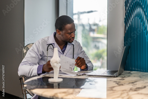 Young doctor man working at office workplace, reading medical record, paper document at laptop, reviewing patient history, sitting at table with laptop computer, working in clinic office