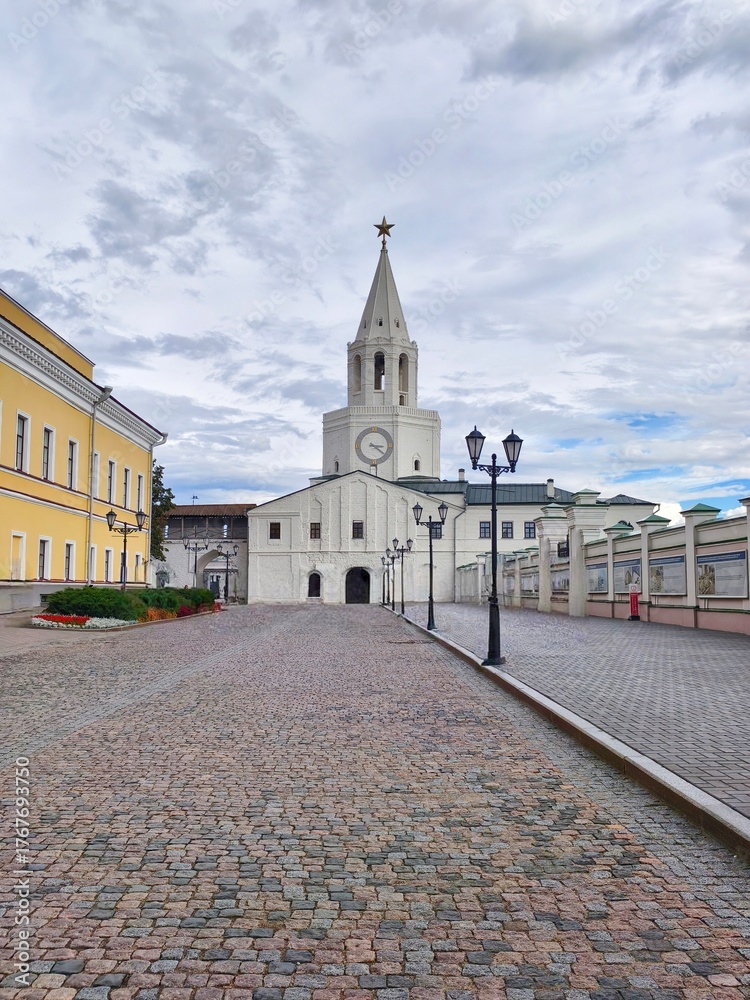 Obraz premium The Spasskaya Tower, main gate of the Kazan Kremlin,a white tower with a clock on it and a tall pointed spire topped with a star and the walkway in front with old-style street lamps lining the sides