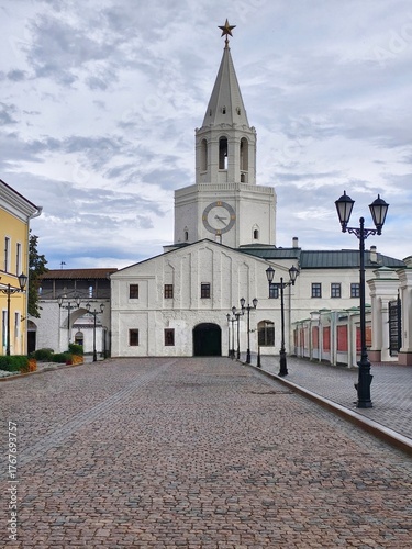 The Spasskaya Tower, main gate of the Kazan Kremlin,a white tower with a clock on it and a tall pointed spire topped with a star and the walkway in front with old-style street lamps lining the sides 