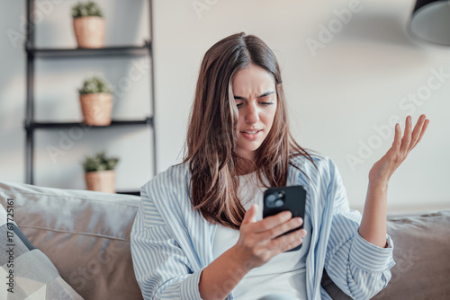 A young woman sits on the sofa, frustrated and upset while using her smartphone. Slow connection, spam, and technical issues leave her stressed and confused at home.