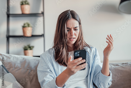 A young woman sits on the sofa, frustrated and upset while using her smartphone. Slow connection, spam, and technical issues leave her stressed and confused at home.