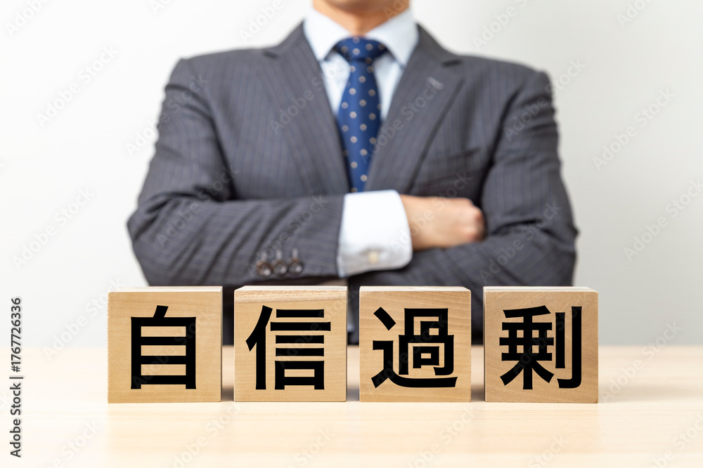 Obraz premium 「自信過剰」と書かれたブロックと腕組みをするビジネスマン