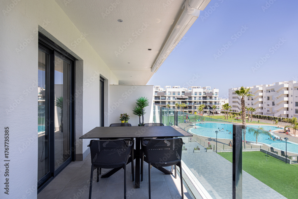 Obraz premium Outdoor dining area on modern apartment balcony with pool view, palm trees, and contemporary buildings under clear sunny sky