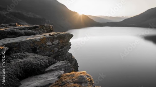 Serene Mountain Lake at Sunrise with Jagged Rocks in Foreground and Golden Sunburst Rays Over Calm Water and Misty Hills