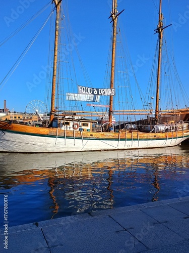 Classic wooden sailboat turned into a floating restaurant, shining under bright sunlight with blue water reflections. Nautical lifestyle and seaside leisure mood.с