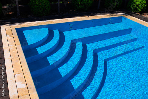 Modern pool with clear blue water and elegant geometric steps casting shadows. Sunlight reflects off the ripples, surrounded by stone edging and tree shadows