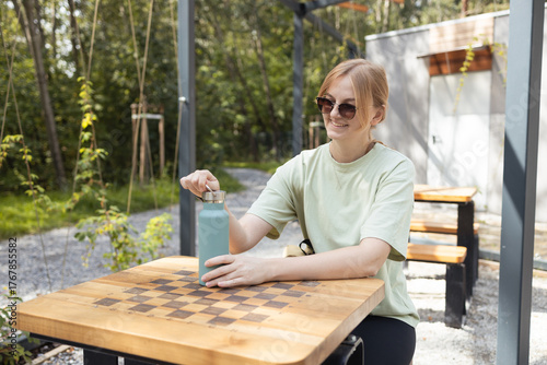 Young woman with sunglasses sitting at a wooden picnic table with a chessboard pattern, opening a reusable water bottle. Concept of eco lifestyle, outdoor relaxation, hydration, and sustainable living