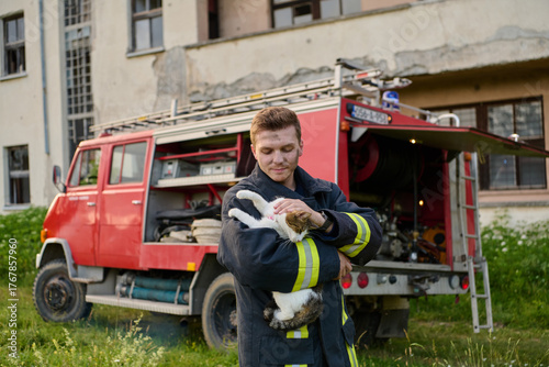 Brave firefighter holding a rescued cat in his arms near a fire truck, showing compassion and care, outdoor scene with a building in the background