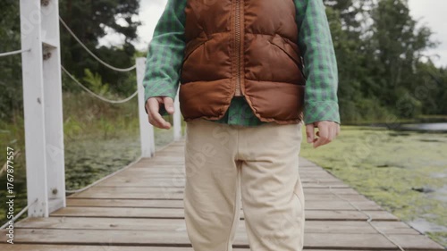 A boy in casual clothes walks calmly on a wooden pier surrounded by greenery. Scene emphasizes healthy lifestyle, balance and connection with nature. High quality 4k footage
