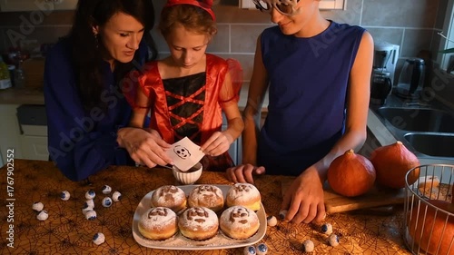 Joyful family in spooky Halloween attire getting ready for a celebration in the kitchen, where a mother and her kids are adding creepy designs and eyeball decorations to donuts 