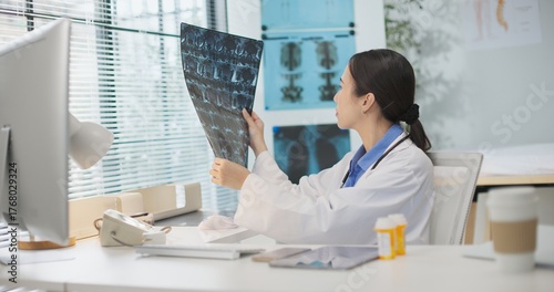 Asian female doctor examines MRI film in clinic, reviewing patient data and medical imaging for diagnosis. Professional healthcare, radiology analysis, spine or neurological check, treatment planning.