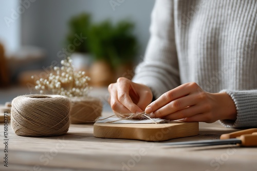 Crafter carefully wraps twine around a small wooden block while sitting at a desk filled with craft supplies and dried flowers to create a handmade gift package with ample copy space.