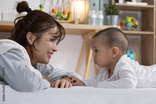 Asian Smiling mother lying on bed facing her baby, gentle eye contact and playful interaction in cozy nursery with toys and soft lighting
