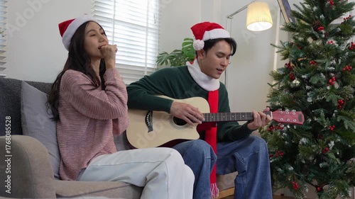 Happy Asian couple celebrating Christmas sharing warmth in winter embrace, exchanging gifts on sofa in cozy home.