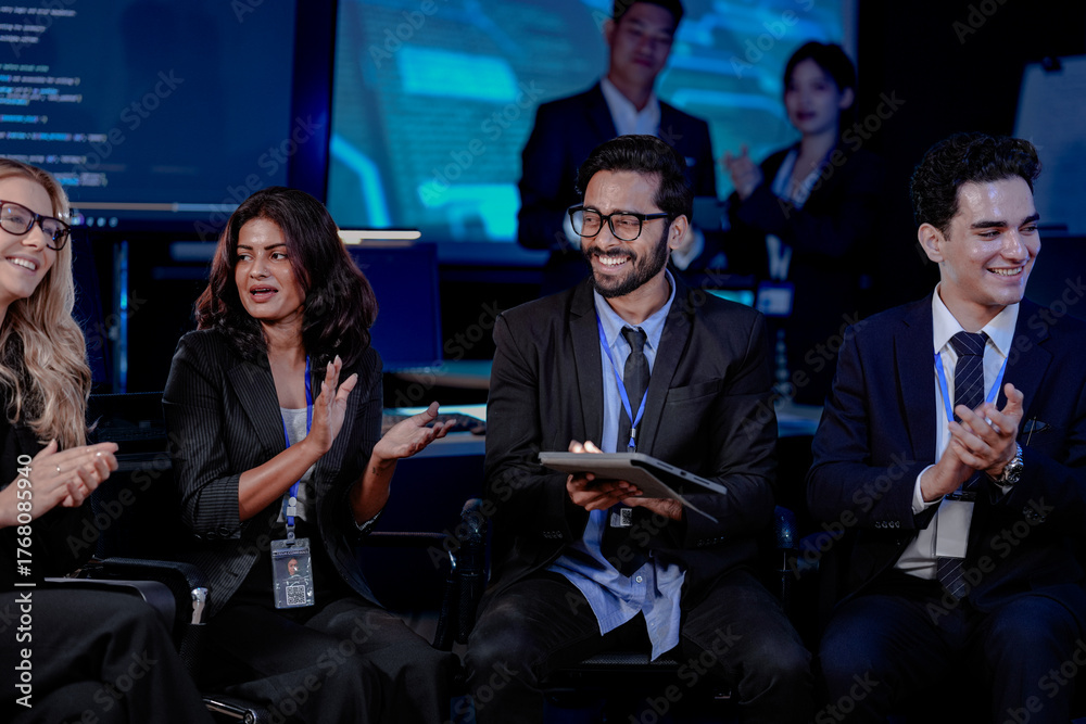 Obraz premium An Indian man with a tablet asks a question during a large team briefing. His diverse colleagues, including Indian and Caucasian women and men, listen in a high-tech conference room.