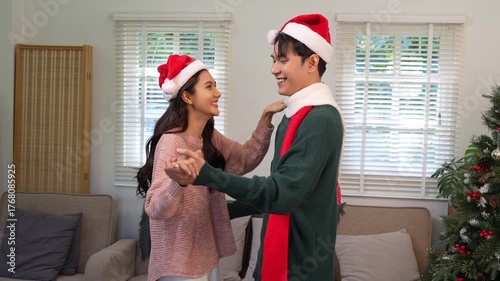 Happy Asian couple celebrating Christmas sharing warmth in winter embrace, exchanging gifts on sofa in cozy home.