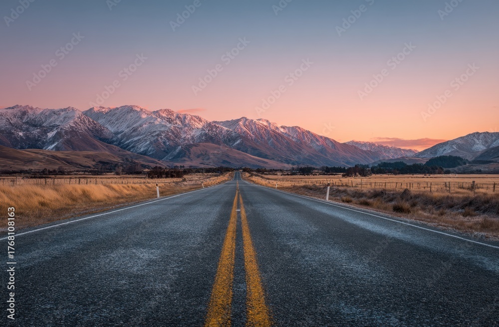 Naklejka premium A long, straight asphalt road stretches into the distance, leading towards snow-capped mountain peaks under a colorful sky.