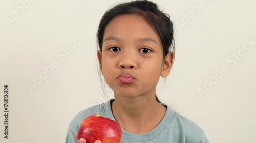 A video shows a girl standing holding a red apple and biting it bit by bit, almost halfway through. With different poses and perspectives, she smiles brightly as she enjoys eating her favourite fruit.