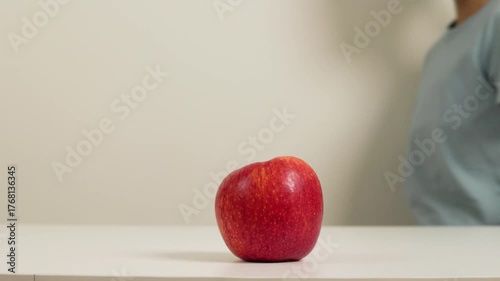 A shiny red apple sits alone on a white table, glowing softly under the light. Suddenly, a curious child walks by, reaches out with a hungry, and quickly grabs the apple before walking away.