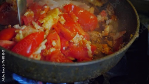 An active and savory mood is captured by a close-up of a spoon stirring scrambled eggs and chopped tomatoes in a hot frying pan. This shot is ideal for depicting home cooking, breakfast