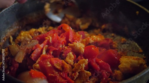 An active and savory mood is captured by a close-up of a spoon stirring scrambled eggs and chopped tomatoes in a hot frying pan. This shot is ideal for depicting home cooking, breakfast
