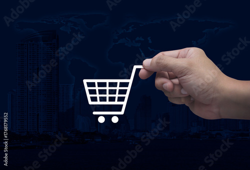 Fototapeta Naklejka Na Ścianę i Meble -  Hand holding shop cart flat icon in fingers over world map, modern city tower and skyscraper, Business shopping online concept, Elements of this image furnished by NASA