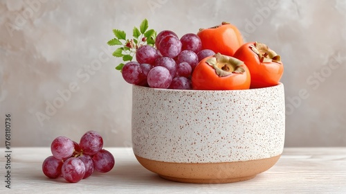 Rustic Still Life Arrangement of Fresh Ripe Persimmons and Purple Grapes in a Textured Ceramic Bowl on a Light Wooden Surface with a Neutral Background