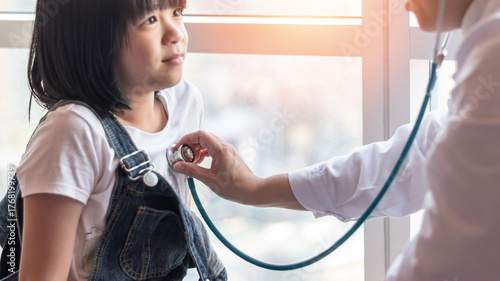 Congenital heart disease defect and pediatric care concept. Asian school child patient with pediatrician doctor examining girl kid health checkup with stethoscope in medical clinic