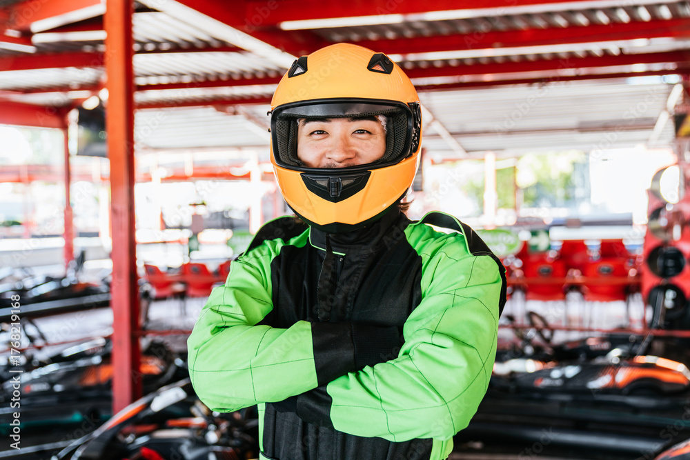 Fototapeta premium Smiling chinese man wearing a driver helmet and racing suit, standing with crossed arms at the karting circuit