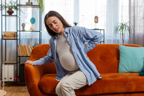 Upset young pregnant woman lady suffering from lower back pain sitting on sofa couch in living room at home. Illness future parent female on couch and feeling sick touching big belly in apartment.