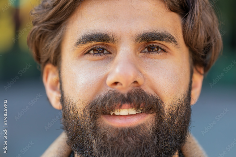 Obraz premium Close-up face portrait of smiling happy Indian man looking at camera, resting, relaxation feel satisfied glad expression of good news outdoors. Bearded young businessman on downtown urban city street