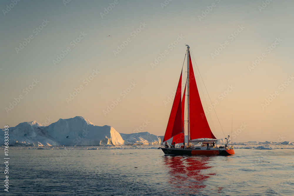 Fototapeta premium A solitary red sailboat courageously navigates through the surreal, glacial landscape of Greenland's Ilulissat Icefjord