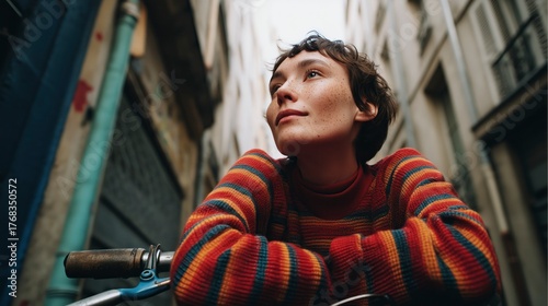 Young person wearing a colorful sweater resting on a bicycle while looking thoughtfully in a narrow urban alleyway during the day
