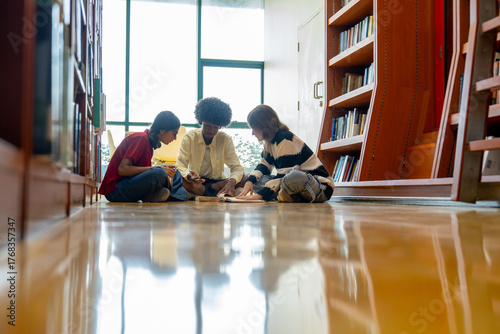 Wide shot of group of teen boy and girls sit on the floor between bookshelves in public library and enjoy to read book and discuss together.