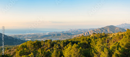 Expansive landscape of Puig Campana region in Spain, featuring lush green pine forests, rolling hills, and distant Mediterranean Sea under a clear sky. Ideal for travel, nature, and tourism.