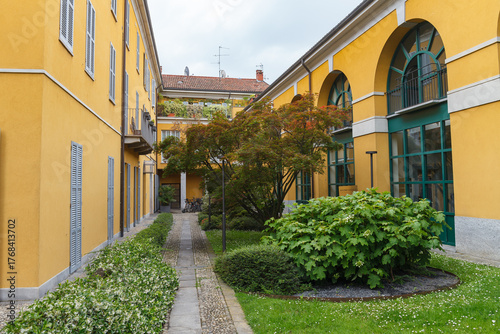 Greenery in the inner yard with pathwalks, historical stone bulding, yellow color, yearly XX century. Monza, Italy.