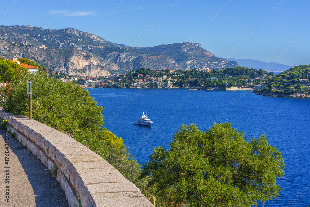 Obraz premium Villefranche-sur-mer, France, French Riviera. Scenic coastal view featuring a yacht sailing in a vibrant blue sea, surrounded by lush greenery and rocky hills, capturing the essence of serenity
