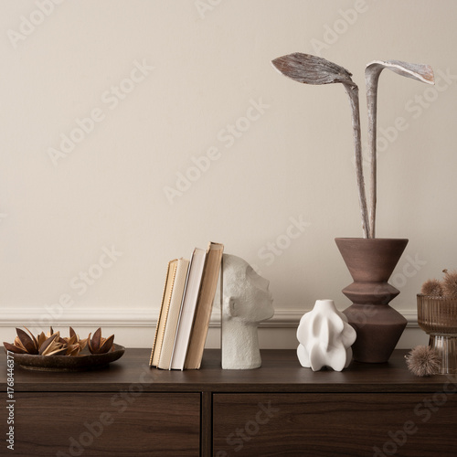 Interior design of aesthetic living room interior with copy space, wooden sideboard, vase with dried flowers, books, modern sculpture, beige wall with stucco and personal accessories. 