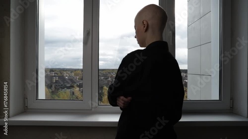Bald woman with cancer standing by window, looking outside at cloudy sky, concept of oncology treatment, hope, and emotional reflection.