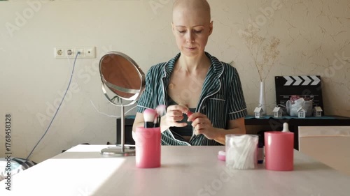 Smiling woman with bald head applies lip gloss at home, completing her makeup routine after chemotherapy