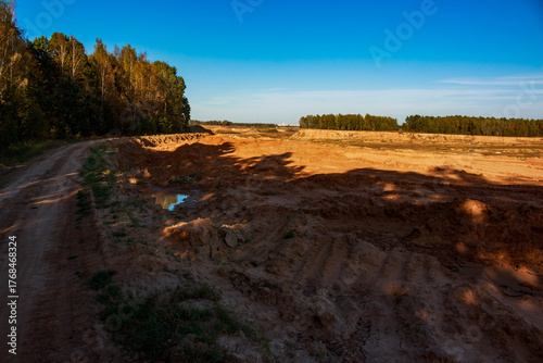 Rugged dirt path through a sprawling sand quarry, bathed in golden light. Autumn foliage lines the distant edges, casting long dramatic shadows