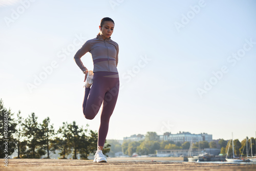 Young adult Caucasian woman stretching leg outdoors on waterfront, preparing for workout, standing on one foot with hand holding ankle, trees and buildings visible in background