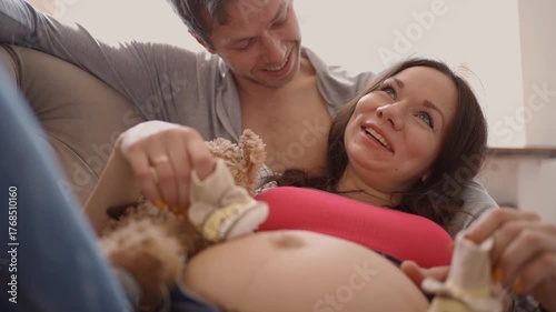 Expecting couple at home lying on sofa, husband gently plays with tiny baby shoes on wifes pregnant belly.