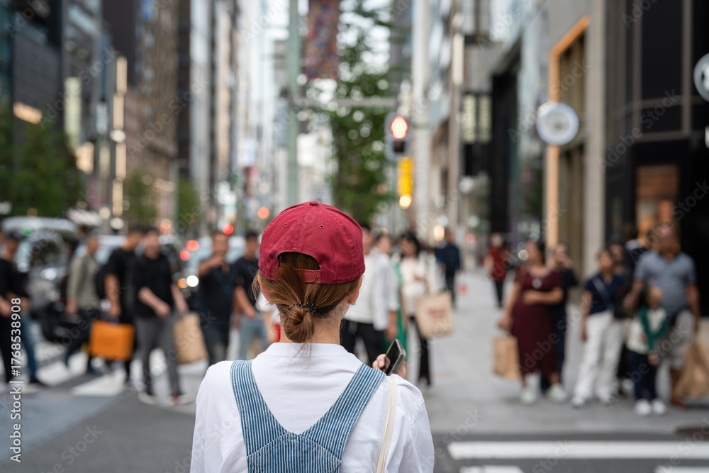 Obraz premium A woman traveler in cozy cloth is waiting to cross the street with crowded of people as blurred background, at downtown shopping area in Tokyo.