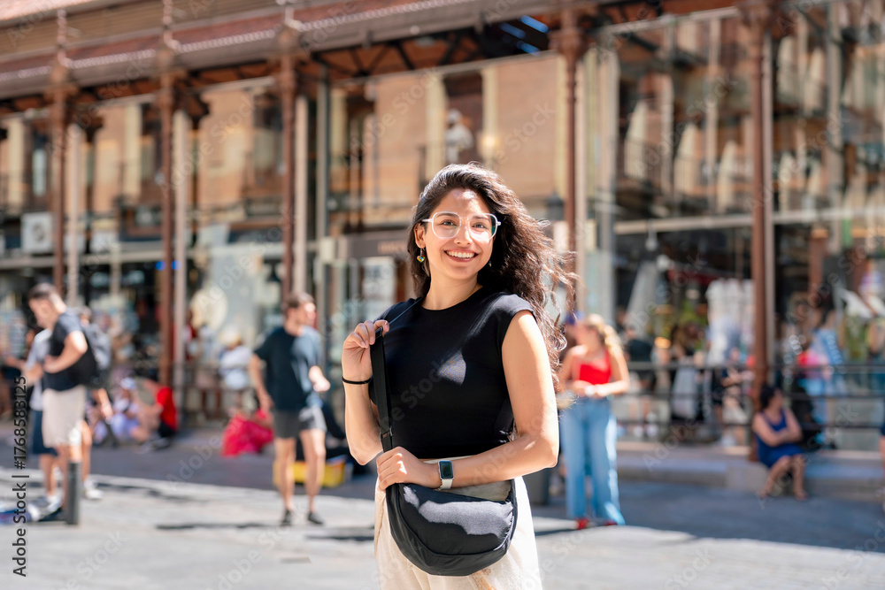 Fototapeta premium Happy young Latin woman smiling outside Mercado de San Miguel in Madrid Spain, wearing casual black and white outfit, famous historic iron market a cultural icon renowned for tapas and gastronomy