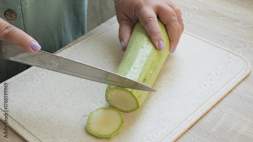 A woman's hand slices green zucchini into rings on a board. Medium shot.