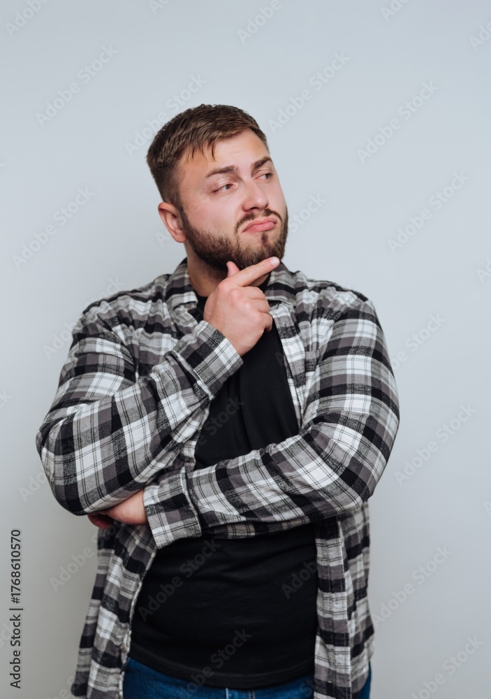 Fototapeta premium Man in plaid shirt pondering with hand on chin against gray background.