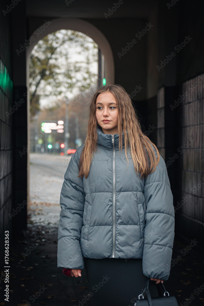 Fototapeta premium Young woman in a gray puffer jacket and black skirt walks through an urban archway on a cloudy autumn day
